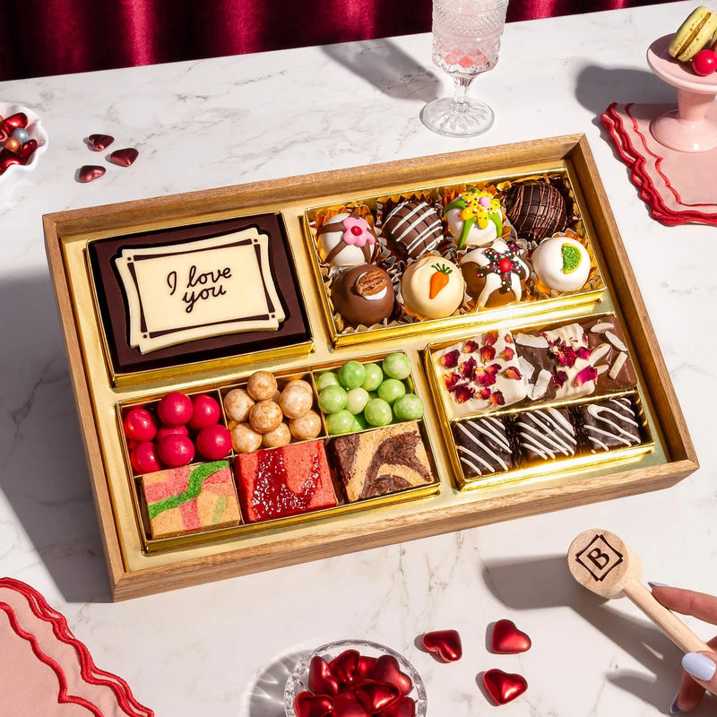 Valentines Day dessert board with "I love you" message, decorated truffles, macarons, candies, and fruit gummies in gold compartments for gifting.