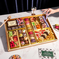 A top-down view of the Thinking of You Dessert Board Large, featuring a reusable acacia wood board filled with over 30 artisan sweets, including macarons, decorated truffles, and gourmet candies in elegant gold compartments. A hand holds a glass nearby, and a small wooden mallet rests next to a central chocolate box engraved with 