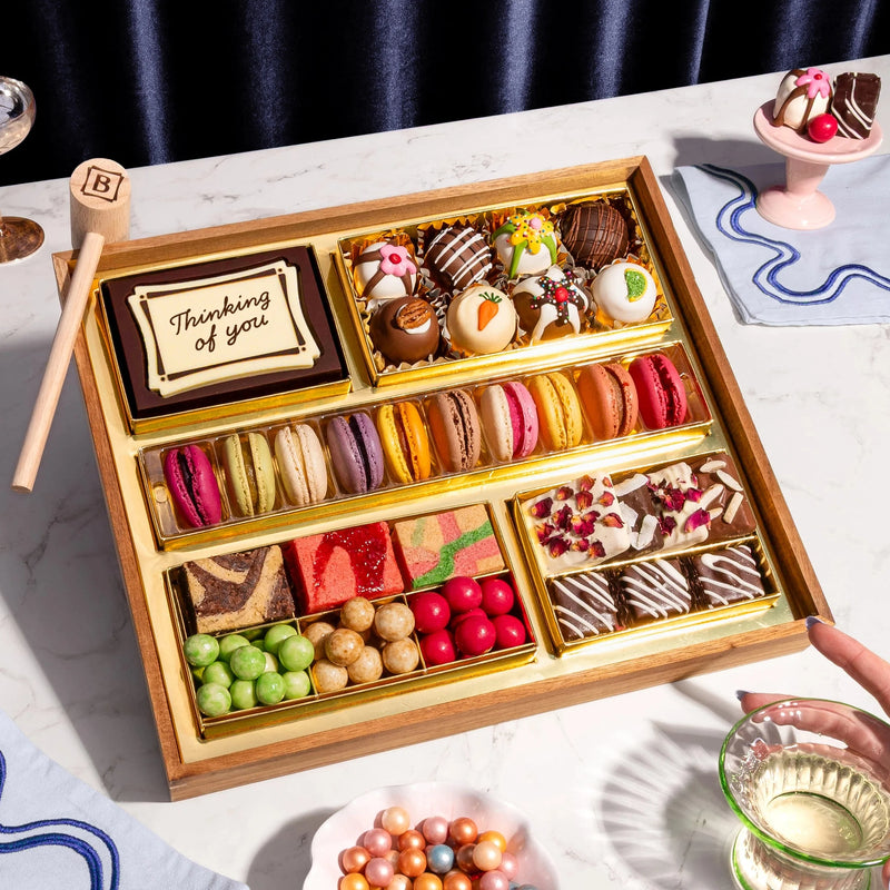 A top-down view of the Thinking of You Dessert Board Medium, featuring a reusable acacia wood board filled with over 30 artisan sweets, including macarons, decorated truffles, and gourmet candies in elegant gold compartments. A hand holds a glass nearby, and a small wooden mallet rests next to a central chocolate box engraved with "Thinking of You."