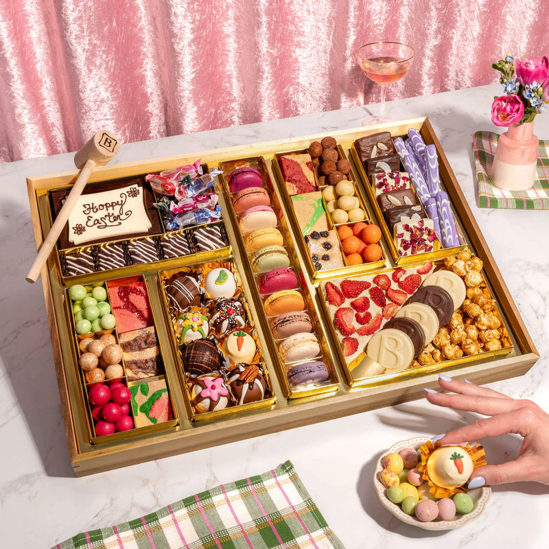 A large Easter dessert charcuterie board with macarons, chocolate-covered strawberries, truffles, carrot-shaped candies, caramel corn, and a 'Happy Easter' cookie with a chocolate mallet.