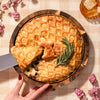 An Easter baked brie charcuterie centerpiece with a golden lattice crust, a slice lifted to reveal melted brie, garnished with rosemary alongside honeycomb and grapes.