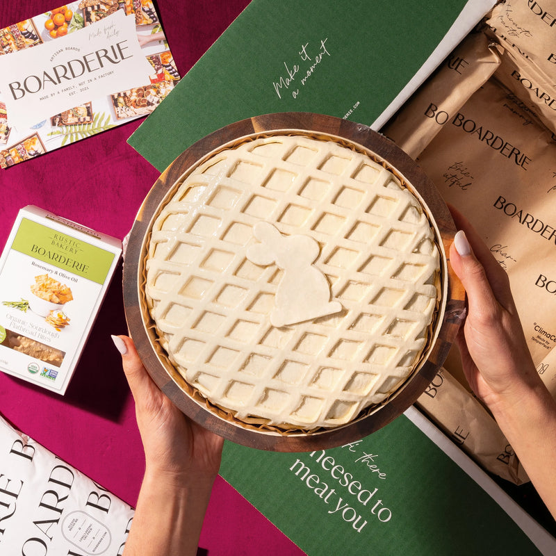 Hands holding a baked brie wheel with a lattice pastry crust and heart cutout, presented over Boarderie gift packaging.