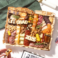 A large graduation charcuterie board topped with 'GRAD' and '2026' cheese letters, cured meats, cheeses, nuts, and dried fruits.