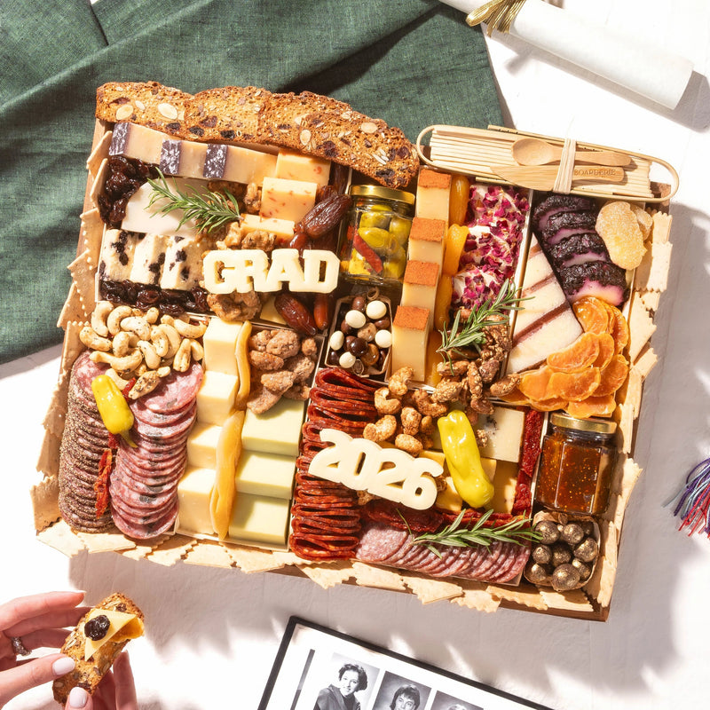 A large graduation charcuterie board topped with 'GRAD' and '2026' cheese letters, cured meats, cheeses, nuts, and dried fruits.