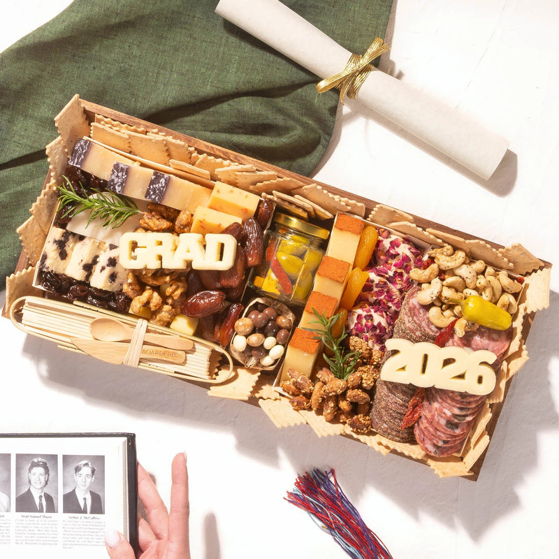 A graduation party charcuterie board with 'GRAD' and '2026' cheese letters, meats, cheeses, and nuts, styled with a diploma scroll and graduation photos.