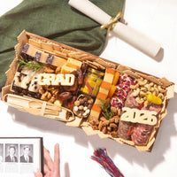 A graduation party charcuterie board with 'GRAD' and '2026' cheese letters, meats, cheeses, and nuts, styled with a diploma scroll and graduation photos.