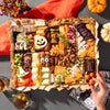 Halloween-themed charcuterie board featuring festive cheeses shaped like a pumpkin and bat, surrounded by assorted snacks and decorations.