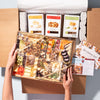 Hands holding a gourmet charcuterie gift box with cheeses, nuts, and crisps, packaged with artisan snacks.