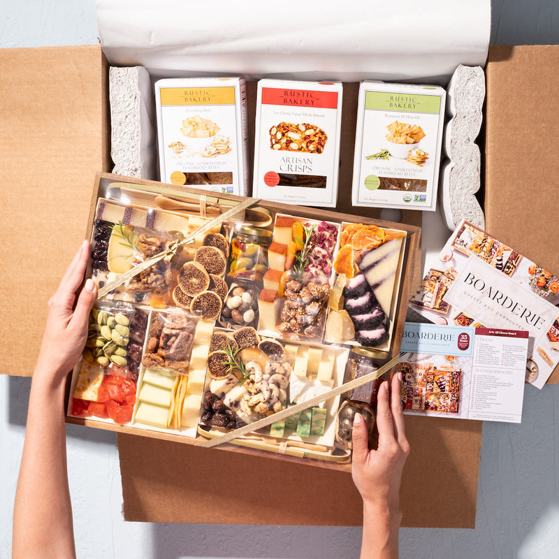 Hands holding a gourmet charcuterie gift box with cheeses, nuts, and crisps, packaged with artisan snacks.