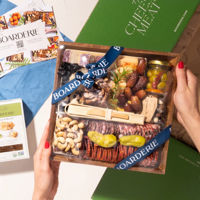 Artisan cheese & charcuterie boards with gourmet meats, cheeses, nuts, dried fruits, olives, and rosemary garnish in gift packaging.
