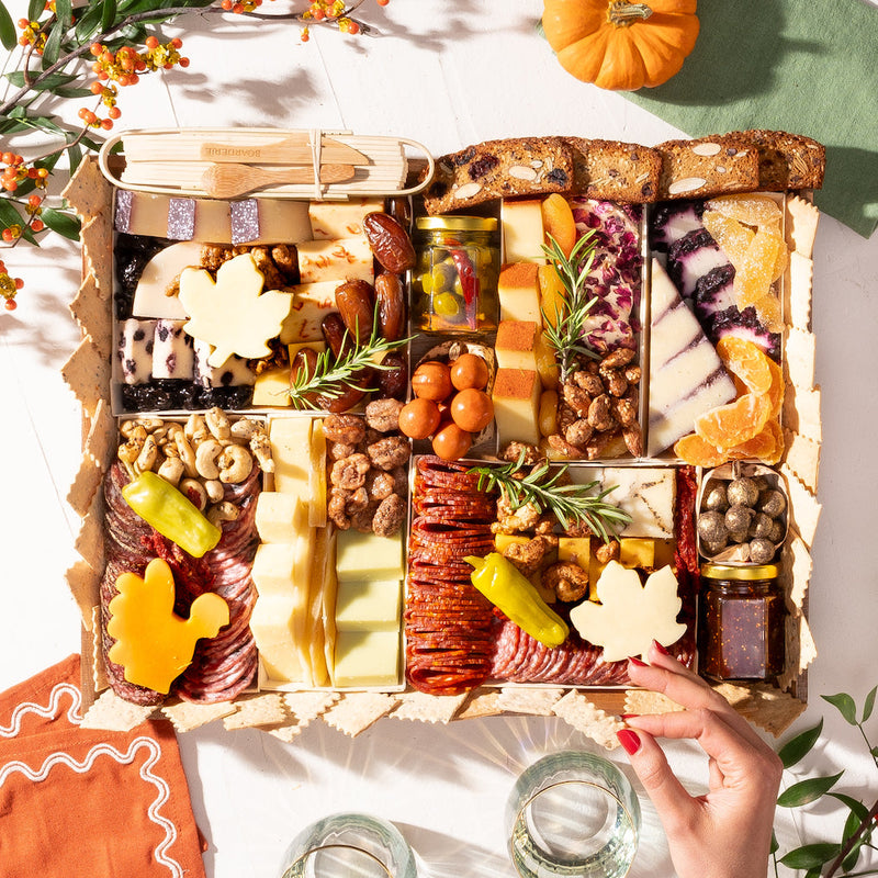 A large Thanksgiving cheese and meat board with maple-leaf and turkey shaped cheeses, cranberry cheese, cured meats, olives, nuts, and dried fruits, styled with a pumpkin and autumn branches.