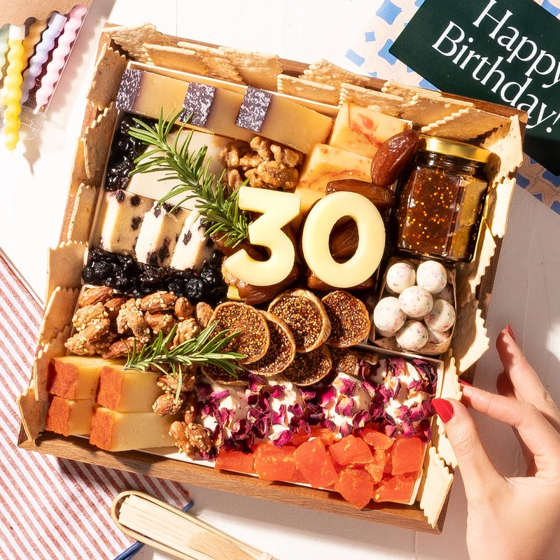 small birthday-themed charcuterie board gift featuring cheeses, nuts, figs, fruit, crackers, & a "30" made of cheese. Fully arranged gourmet birthday gift with 19 artisan cheeses, dried fruits, nuts, chocolates, jam and crackers on a reusable acacia wood board. Includes bamboo utensils, birthday candles, confetti and customizable age numbers in cheese. Option to add personal gift note. Ships free overnight nationwide in insulated packaging with next-day delivery available.