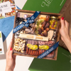 Hands holding a Boarderie charcuterie box featuring an assortment of cheeses, nuts, dried fruits, &  fig spread, wrapped in branded ribbon