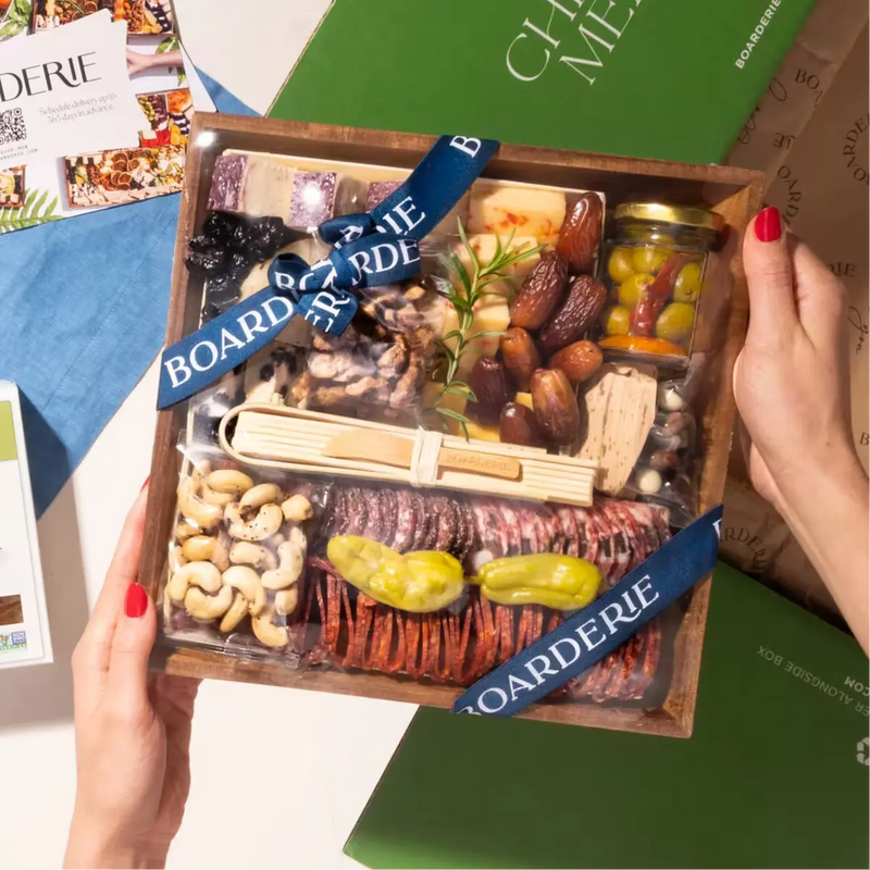 Hands holding a Boarderie charcuterie box featuring an assortment of cheeses, nuts, dried fruits, &  fig spread, wrapped in branded ribbon