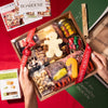 A Christmas gift charcuterie board being unboxed with a gingerbread-man cheese, cured meats, nuts, and crackers wrapped in a red 'Happy Holidays' ribbon.
