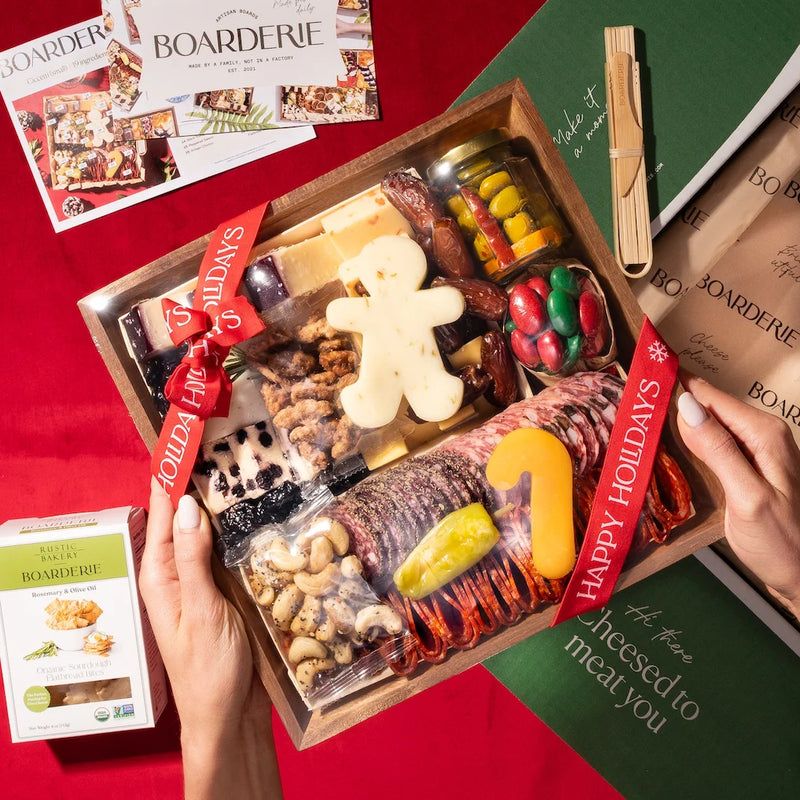 A Christmas gift charcuterie board being unboxed with a gingerbread-man cheese, cured meats, nuts, and crackers wrapped in a red 'Happy Holidays' ribbon.