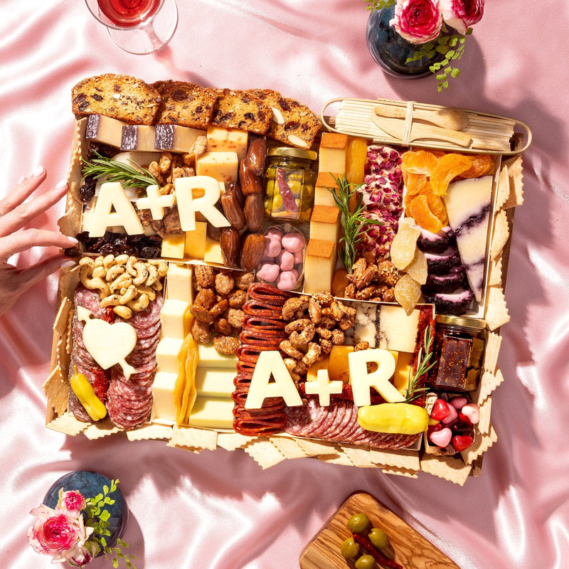 Luxury Valentine’s Day charcuterie board gift by Boarderie featuring artisan cheeses, cured meats, dried fruits, nuts, crackers, gourmet spreads, and customized initials in cheese. Board is on a pink romantic backdrop, styled for nationwide overnight gifting. 