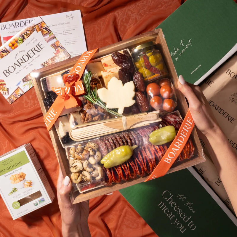 A Thanksgiving charcuterie board being unboxed, featuring a maple leaf cheese, cured meats, olives, nuts, and dates in a Boarderie gift box with an orange ribbon.