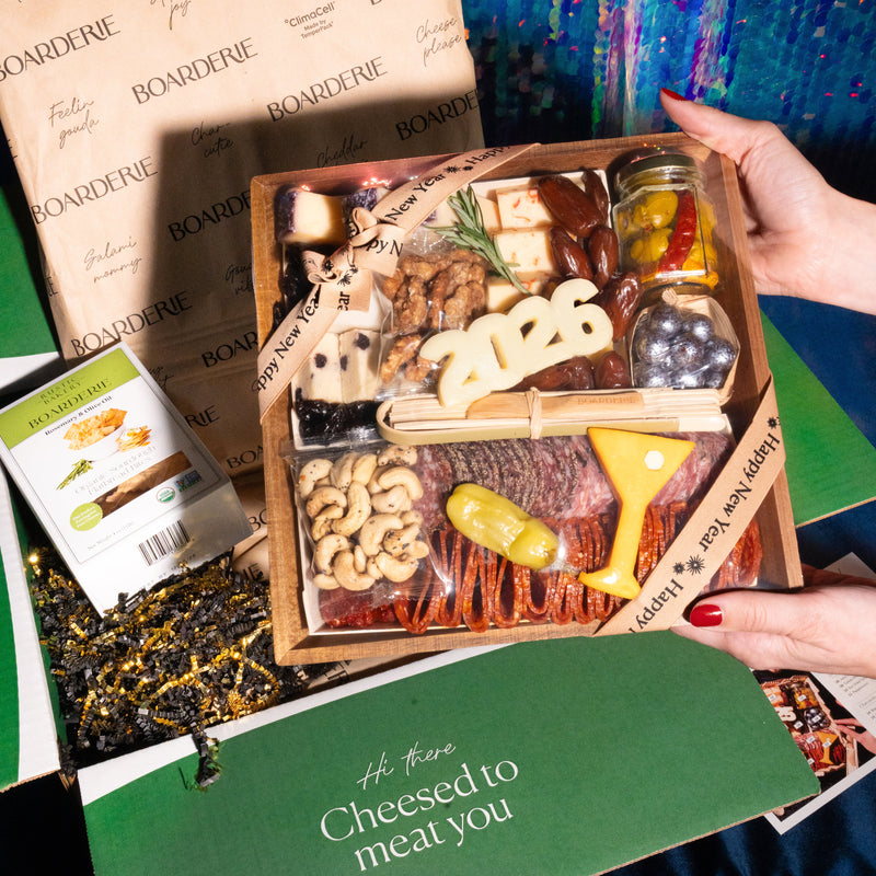 New Year's  charcuterie board gift with "2026" cheese cutout, cured meats, cheeses, nuts, pickled vegetables, serving utensils, and branded Boarderie packaging.