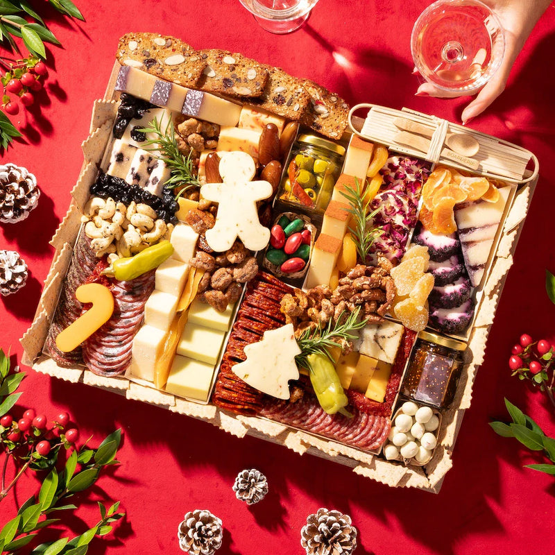 large Festive charcuterie board with holiday-themed cheeses, meats, and nuts on a red background with pinecones & holly
