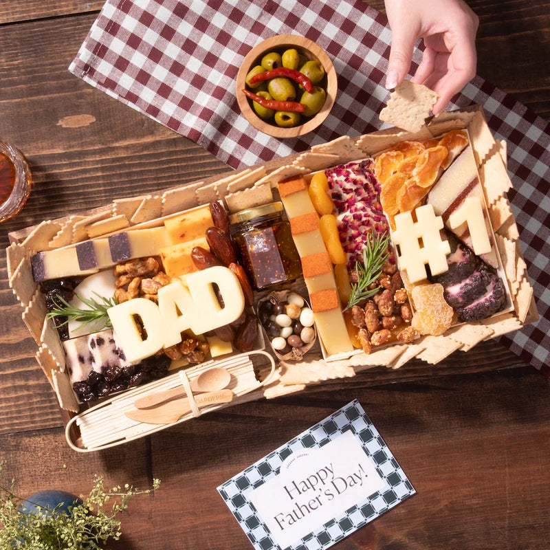 Cheese board for Father's Day featuring artisan cheeses, dried fruits, nuts, olives, jam, and “DAD” and “#1” shaped cheeses, arranged in a gift box with crackers and a wooden utensil set.