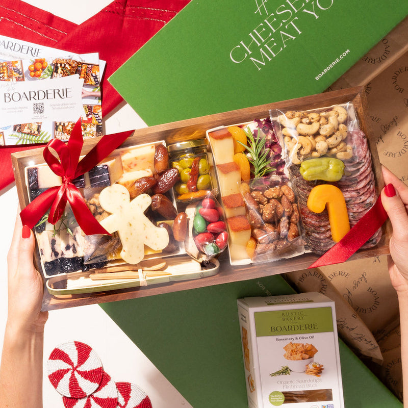 Holiday-themed small cheese and charcuterie board with festive cheese shapes, nuts, dried fruits, and crackers, tied with a red ribbon and boxed with artisan crisps