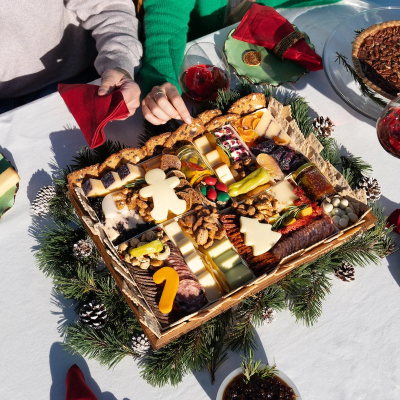 A Christmas themed charcuterie board with candy-cane and gingerbread-man shaped cheeses, cured meats, nuts, and chocolates, set on a holiday table with pine boughs and pinecones.