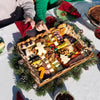 A Christmas themed charcuterie board with candy-cane and gingerbread-man shaped cheeses, cured meats, nuts, and chocolates, set on a holiday table with pine boughs and pinecones.