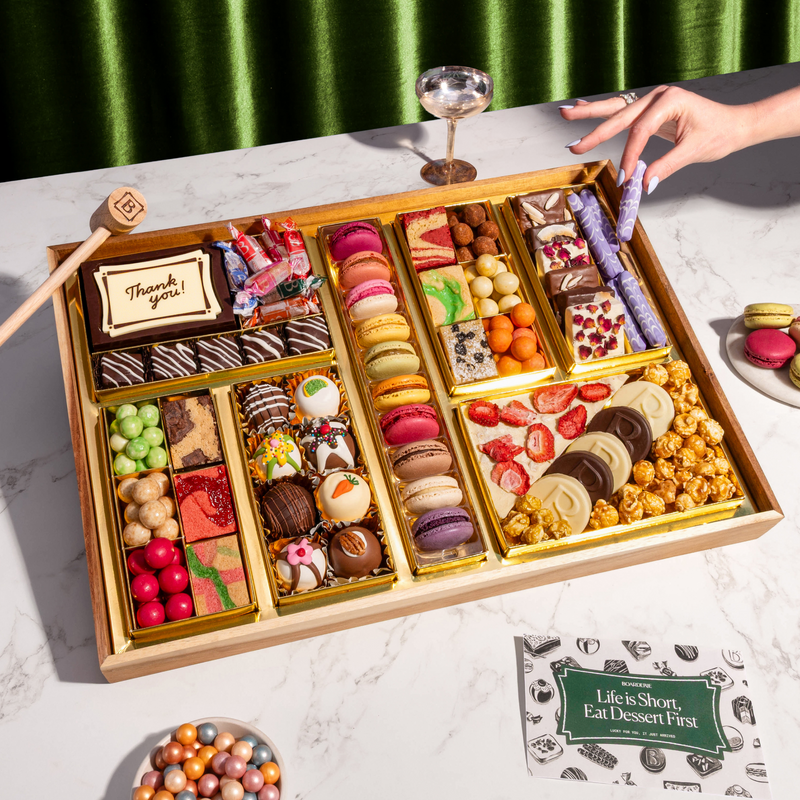 Dessert gift box filled with macarons, decorated chocolate truffles, brownies, caramel popcorn, strawberry bark, candies, and a "Thank you" chocolate plaque in a gold-lined wooden tray on a marble surface
