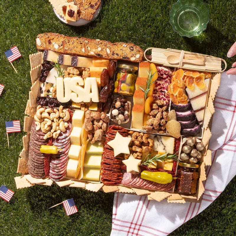 Patriotic charcuterie board with meats, cheeses, nuts, and cheese cutouts spelling “USA” and shaped like stars.