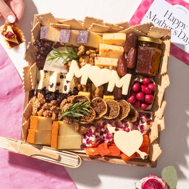 Small Mother's Day cheese board with assorted cheeses, dried fruits, nuts, and "MOM" and heart-shaped cheese cutouts.