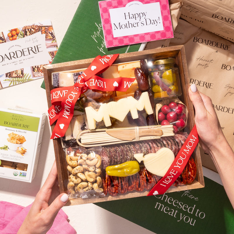 Mother's Day charcuterie box with gourmet meats, cheeses, and festive "MOM" and heart cheese cutouts in gift packaging.