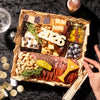 Wooden New Years charcuterie board displaying assorted cheeses, cured salami slices, roasted cashews, marinated olives, berry jam, dark chocolate pieces, and decorative 2026 numeral with fresh rosemary sprigs.
