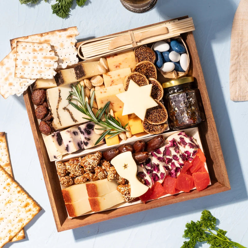 Passover charcuterie board with matzo, dried fruits, nuts, herb-garnished cheeses, fig jam, and star and bow-shaped cheese accents.