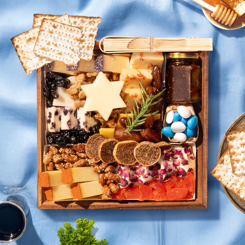 Small Passover cheese board with a Star of David cheese cutout, blueberry cheese, sliced dried figs, rose petal cheese, candied nuts, dried apricots, fig jam, and blue and white candies alongside matzo on a blue linen surface