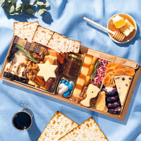 Medium Passover cheese board featuring matzo, a Star of David cheese cutout, artisan cheeses, candied walnuts, dates, fig jam, dried fruit, and blue and white candies on a blue linen backdrop