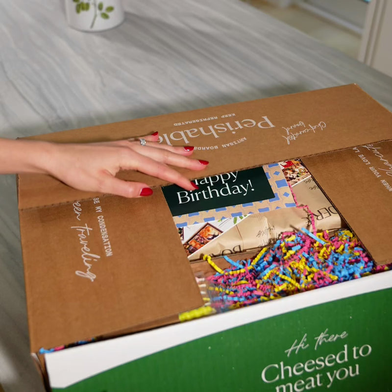 charcuterie board birthday gift video of unboxing the fresh board in a beautiful kitchen. Charcuterie board includes candles and crackers. Charcuterie board is unwrapped and candles are lit and blown out.