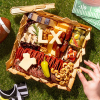 Super Bowl-themed charcuterie board with cheeses, meats, crackers, nuts, and football-inspired cheese cutouts on a grass picnic setting.