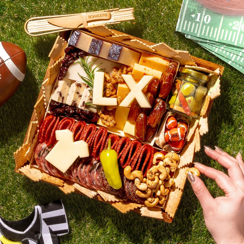 Super Bowl-themed charcuterie board with cheeses, meats, crackers, nuts, and football-inspired cheese cutouts on a grass picnic setting.