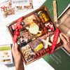 Valentine's Day wooden charcuterie board gift with cured meats, cheeses, nuts, pickles, and white chocolate hearts tied with red ribbons.