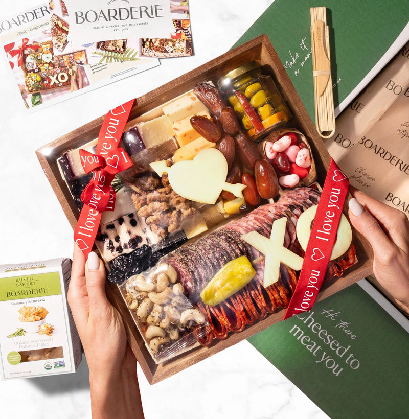 Valentine's Day wooden charcuterie board gift with cured meats, cheeses, nuts, pickles, and white chocolate hearts tied with red ribbons.